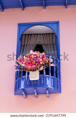 Vertical photo of antique Latin American architecture of a window decorated with a basket of roses and flowers, painted in dark blue and pastel pink.