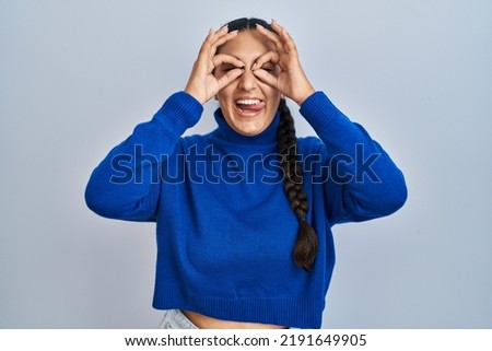 Young hispanic woman standing over isolated background doing ok gesture like binoculars sticking tongue out, eyes looking through fingers. crazy expression. 