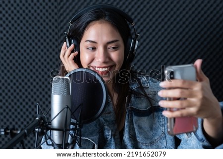 Happy cheerful pretty smiling of portrait young Asian woman vocalist Wearing Headphones taking a selfie with smartphone while  recording a song front of microphone in a professional studio