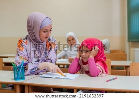 Female hijab Muslim teacher helps confused and stressed school child to finish the lesson in the classroom.	
