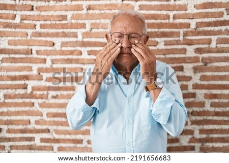 Senior man with grey hair standing over bricks wall rubbing eyes for fatigue and headache, sleepy and tired expression. vision problem 