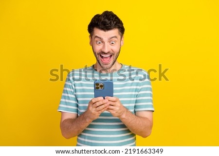 Photo of funny ecstatic handsome guy dressed striped t-shirt look on smartphone read message impressed sale on yellow color background