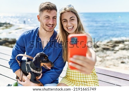 Young caucasian couple make selfie by the smartphone sitting on the bench with dog at the beach.
