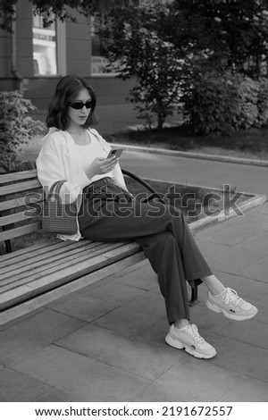 Beautiful and tall brunette is sitting on a bench in the park. Stylish look. Business woman lifestyle. White shirt, black pants, small bag. Black sunglasses. Summer city. Black and white photo.