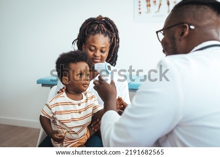 Smiling caring male pediatrician talk consult small boy patient at consultation in hospital with mother. Doctor measuring temperature of boy with infrared thermometer at hospital 