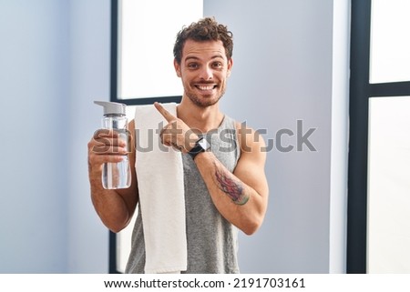Young hispanic man wearing sportswear drinking water cheerful with a smile on face pointing with hand and finger up to the side with happy and natural expression 