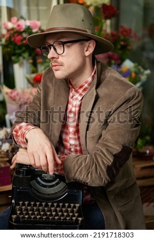 Cute male person in eyeglasses and hat sitting with old typewriter in flower store. Poet or writer working concept. High quality vertical image