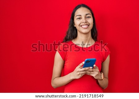 Young brazilian woman using smartphone over red background with a happy and cool smile on face. lucky person. 