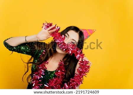 Young beautiful girl 20-25s in a party hat and New Year's tinsel in her hands poses and looks at the camera on a yellow isolated background. Makes an impromptu mustache from New Year's tinsel. Royalty-Free Stock Photo #2191724609