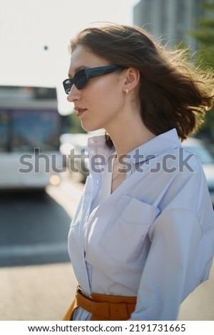 Beautiful modern woman walking down the street. Pale blue clothes. Knitted bag. Orange belt with basque. Brown hair. Sunglasses. Slim body, long legs. Daily life. Work and leisure. City landscape.