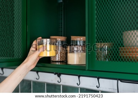 storage grains and beans in jar on shelf in metal green cabinet at kitchen and female hand taking glass container with wheat for prepare food Royalty-Free Stock Photo #2191750423