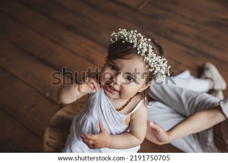 Portrait of pretty one year girl in white summer dress, flower crown and with eco beads sitting in the studio near mom's legs. Eco style concept Royalty-Free Stock Photo #2191750705
