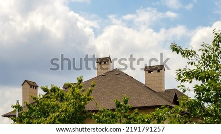 Roof made of brown bituminous tiles, roofing shingles and three chimneys against blue sky with white clouds. Trees in the foreground. 16 by 9, web banner. Royalty-Free Stock Photo #2191791257