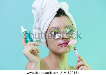  Horizontal photo, a woman with radiant skin on a blue background in a towel on her head and body and iridescent glasses and patches on her lips takes care of her face with serum