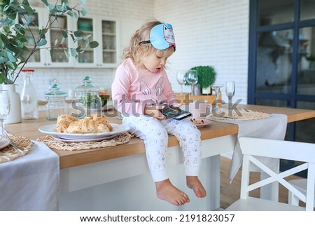 Adorable toddler eating fresh croissants and using phone. Girl dressed in pajama, sleep mask, watching cartoons while eating. Digital technologies, kids education concept. Child addiction from gadgets