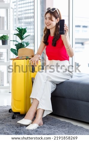 Asian pretty cheerful happy female traveler in casual summer outfit with sunglasses and hat sitting smiling with yellow trolley luggage using smartphone browsing surfing internet booking hotel online.