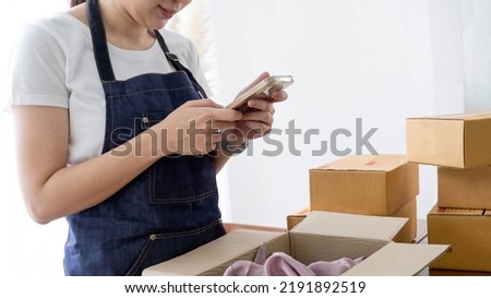 Hands of Asian woman entrepreneur holding smartphone with cardboard boxes, computer laptop, online selling equipments. Packing, business and technology concept. Top view, copy space
