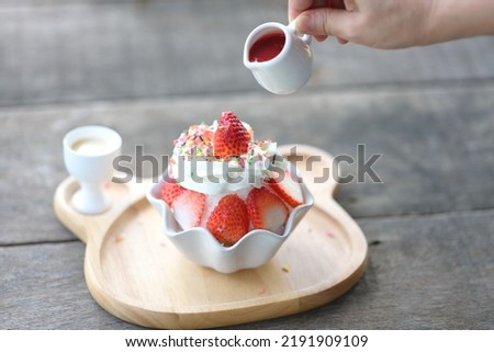 Woman's hands pouring strawberry sauce in a small white bowl over milk cream in shaved ice and vanilla ice cream or bingsu (traditional Korean cold fruit dessert). A popular traditional summer dessert