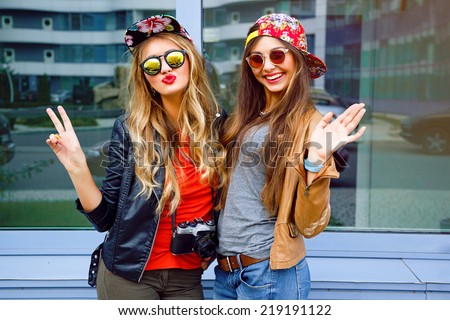 Bright stylish lifestyle urban portrait of two pretty best friends girls posing at leather jackets bright swag hats and sunglasses. Having faun, send you kiss and say hello.