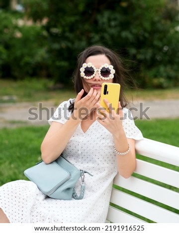 Caucasian brunette woman in trendy sunwear makes selfie sitting on bench in city park on cloudy summer day. Attractive girl in white sundress and optical round sunglasses poses for selfie outdoors