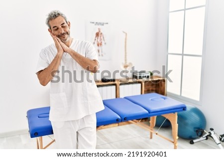 Middle age hispanic therapist man working at pain recovery clinic sleeping tired dreaming and posing with hands together while smiling with closed eyes. 