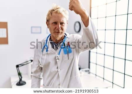 Young blond man wearing doctor uniform and stethoscope at clinic angry and mad raising fist frustrated and furious while shouting with anger. rage and aggressive concept.  Royalty-Free Stock Photo #2191930513