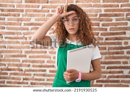 Young caucasian woman holding art notebook making fun of people with fingers on forehead doing loser gesture mocking and insulting. 