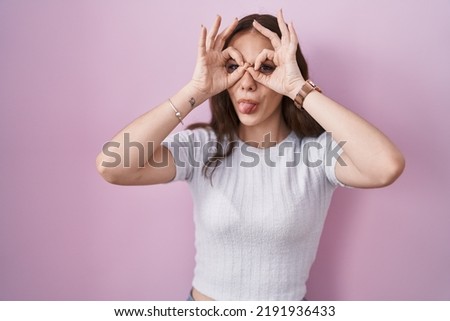 Young hispanic girl standing over pink background doing ok gesture like binoculars sticking tongue out, eyes looking through fingers.   expression. 