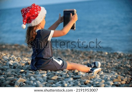 a little girl in a blue dress and a red santa claus hat sits on the seashore in the sunset and watches cartoons on a tablet, plays and chats online with friends