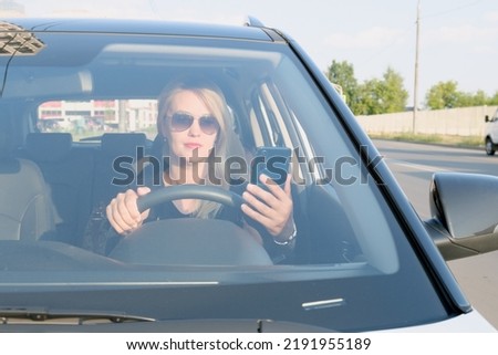 Young attractive woman with sunglasses sits at the wheel of a car with a smartphone in her hand. Beautiful blonde woman using the phone while driving. Shot through windshield. Royalty-Free Stock Photo #2191955189
