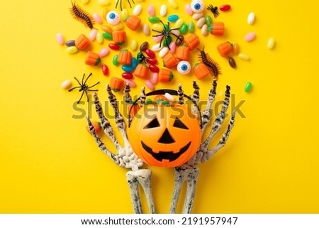 Halloween concept. Top view photo of skeleton hands holding pumpkin basket with candies eyes centipedes and spiders on isolated yellow background