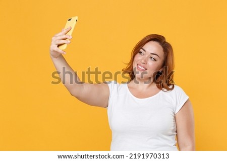 Smiling beautiful young redhead plus size body positive female woman girl 20s in white casual t-shirt posing doing selfie shot on mobile phone isolated on yellow color wall background studio portrait
