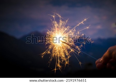 Hand holding a sparkler, or bengal fire stick, burning in outdoor setting, mountain landscape, at dusk. Holidays or magic background or wallpaper. Royalty-Free Stock Photo #2191976651
