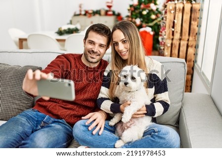 Young hispanic couple making selfie by the smartphone sitting on the sofa with dog at home.