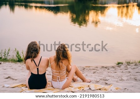 Rear view of two girls in bikini sitting on a beach. Girlfriends at picnic on a lake or river coast, summer leisure on a nature, female friendship.