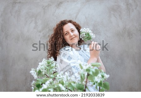 Ukrainian woman with curly hair in white embroidered shirt. Defocused focus in foreground on blossoming branches of apple tree against the background of a concrete wall. Independence Day of Ukraine.