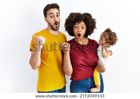 Interracial young family of black mother and hispanic father with daughter surprised pointing with hand finger to the side, open mouth amazed expression. 