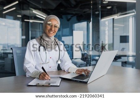 Portrait of female doctor in hijab, muslim working in modern office inside clinic, smiling and looking at camera, filling documents, using laptop
