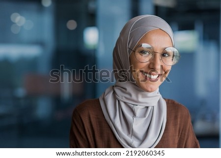 Close up photo portrait of beautiful young muslim woman, woman in hijab and glasses smiling and looking at camera, businesswoman working inside modern office building