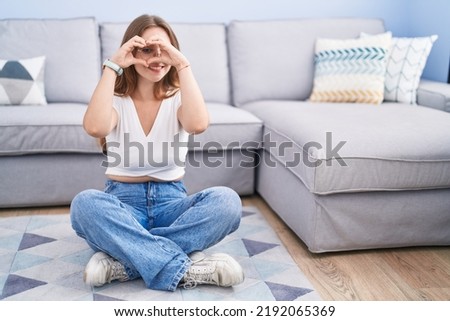 Young caucasian woman sitting on the floor at the living room doing heart shape with hand and fingers smiling looking through sign 