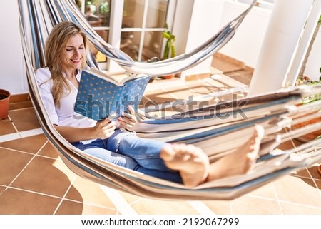 Young blonde woman reading book lying on hammock at terrace.