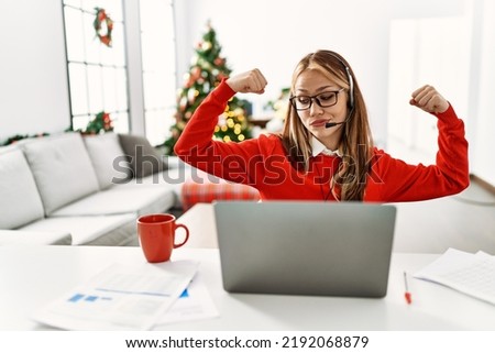 Young caucasian girl sitting on the table working using laptop by christmas tree showing arms muscles smiling proud. fitness concept. 