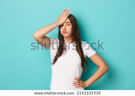 Young tired woman in white t-shirt, sighing and making facepalm to express annoyance, feeling fed up and tensed, standing over blue background Royalty-Free Stock Photo #2192101939