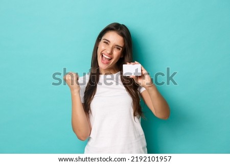 Image of satisfied ecstatic woman, wearing white t-shirt, rejoicing and showing credit card, standing pleased over blue background Royalty-Free Stock Photo #2192101957