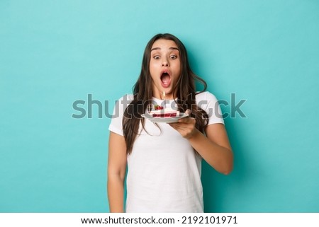 Image of excited birthday girl in white t-shirt, making wish and blowing candle on cake, celebrating bday over blue background