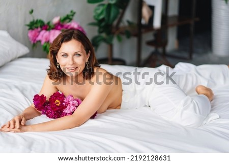attractive brunette topless woman with a bouquet of peonies on the bed. women's happiness and psychological health. fragrances for home and body. beautiful bed linen made of natural fabrics. comfort