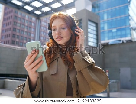 Teen redhead girl standing on urban street skyscrapers background wearing headphones using smartphone listening to music, audio book, travel guide or podcast while walking in big city. Royalty-Free Stock Photo #2192133825