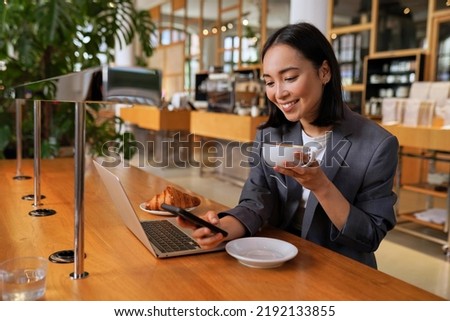 Young Asian business woman wearing suit drinking coffee using smartphone in cafe. Happy smiling female professional working holding mobile phone using smartphone texting messages on cellphone. Royalty-Free Stock Photo #2192133855