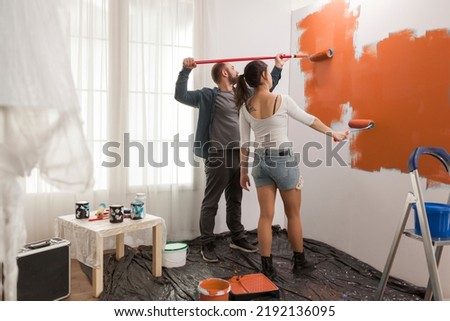 Happy couple painting wall with orange paint, redecorating apartment with new color. House renovation and improvement with paintbrush, manual roller, renovating living room walls. Royalty-Free Stock Photo #2192136095