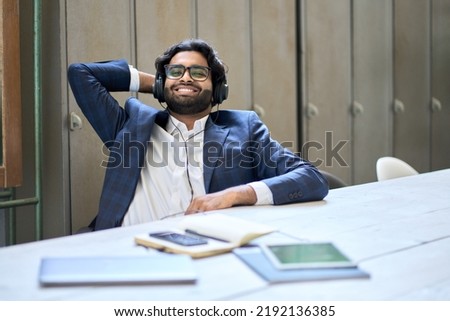 Young happy satisfied indian business man employee or executive wearing suit and headphones sitting at work desk listening to music relaxing after work done, having pleasant break at workplace. Royalty-Free Stock Photo #2192136385